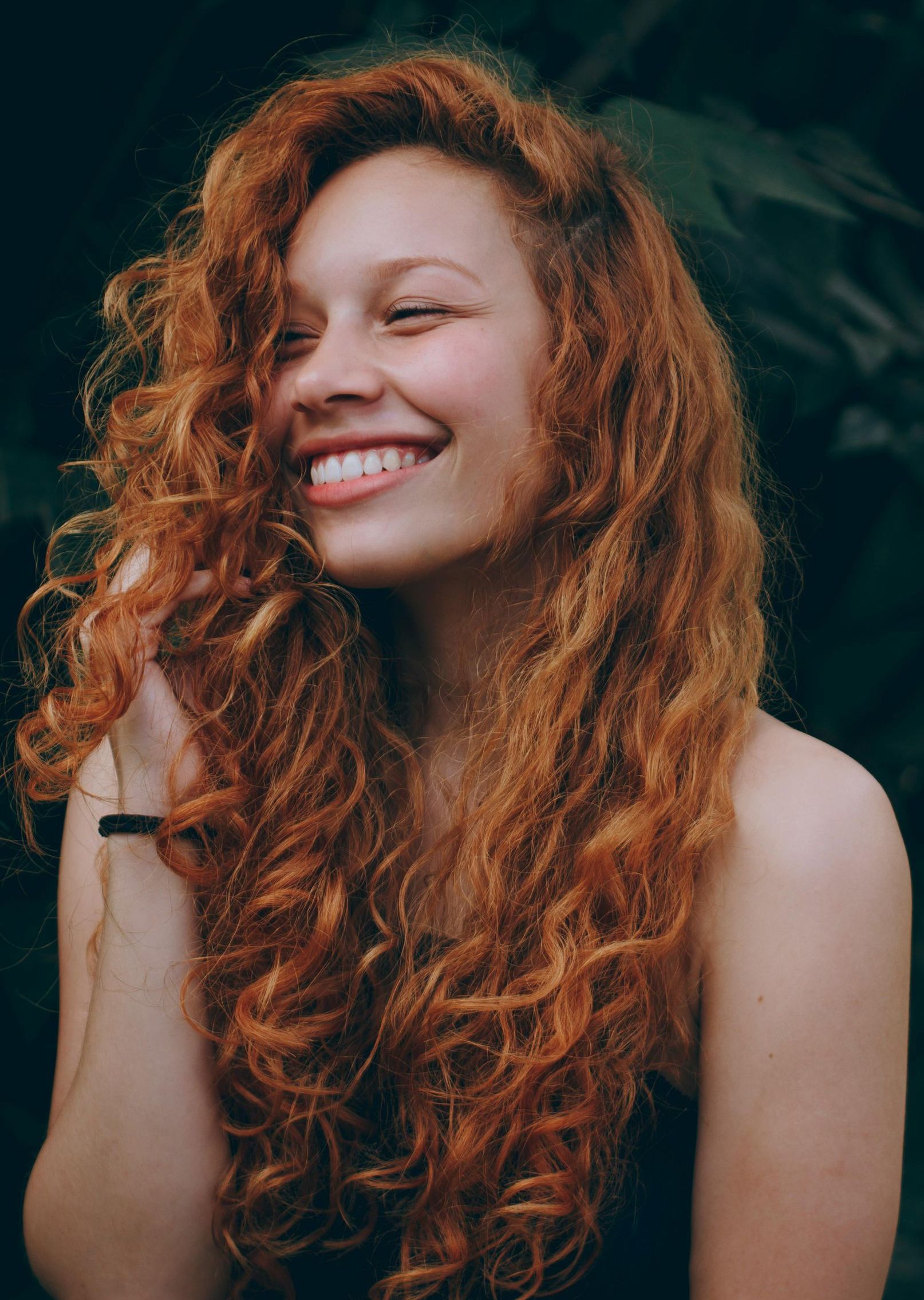 A woman with long red curly hair is smiling and holding her hair.