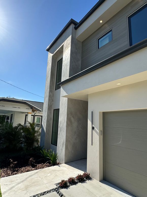 A gray stucco wall with two small windows, a white door, and an electrical box mounted on the exterior.