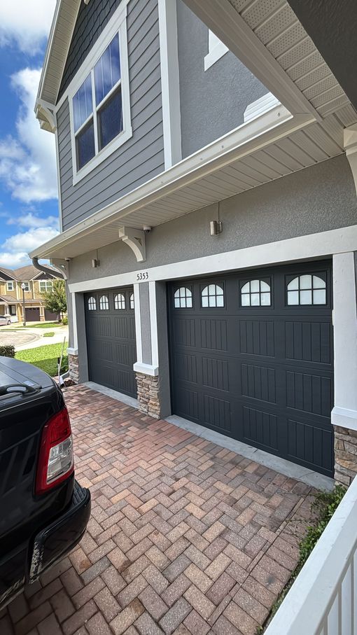 A two-story gray house with two dark gray garage doors, a brick driveway, and a black vehicle parked in the foreground.