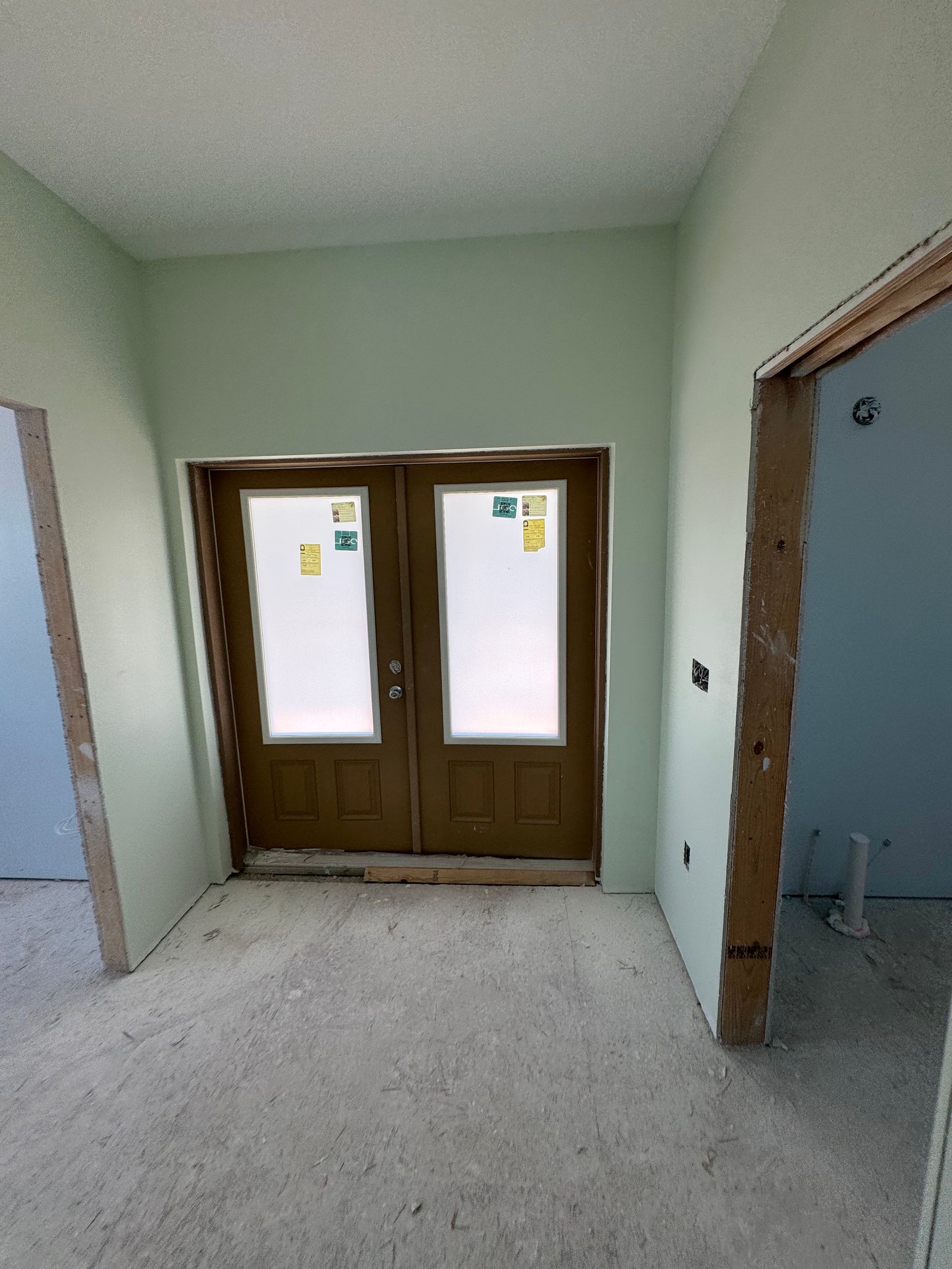 A view of a doorway with double doors in a room undergoing construction, featuring light green walls and unfinished floors.