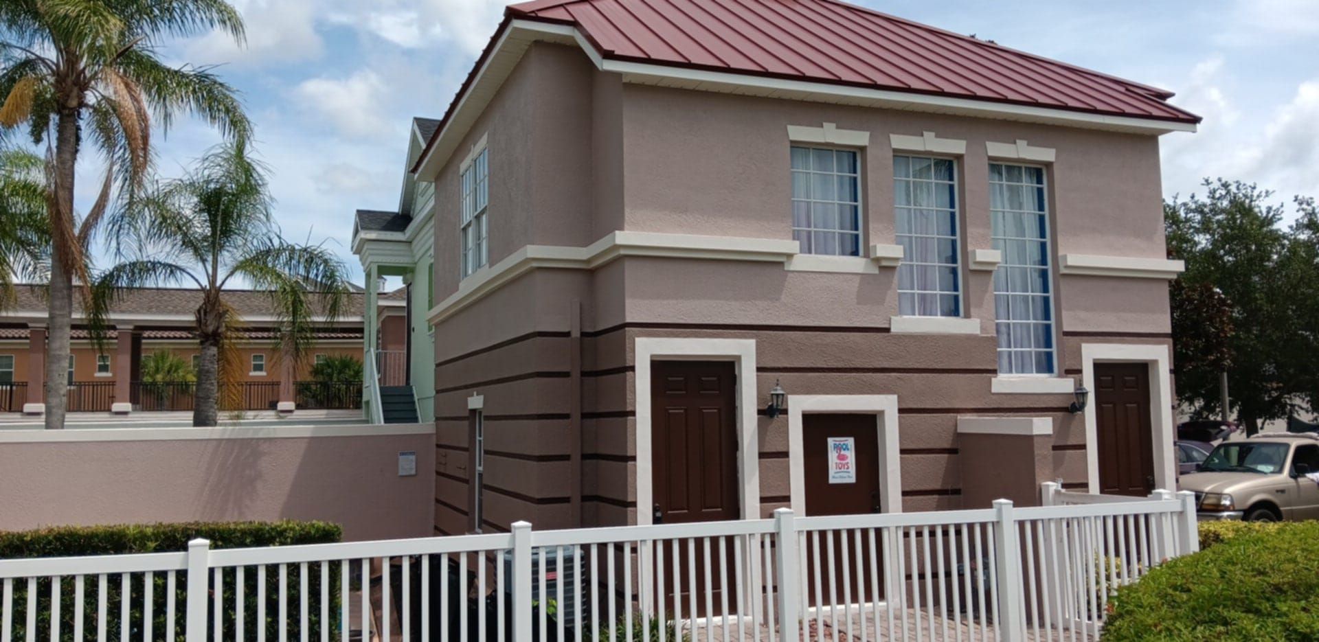 A two-story tan building with a reddish roof, brown doors, and white fencing, situated in a sunny, landscaped setting.