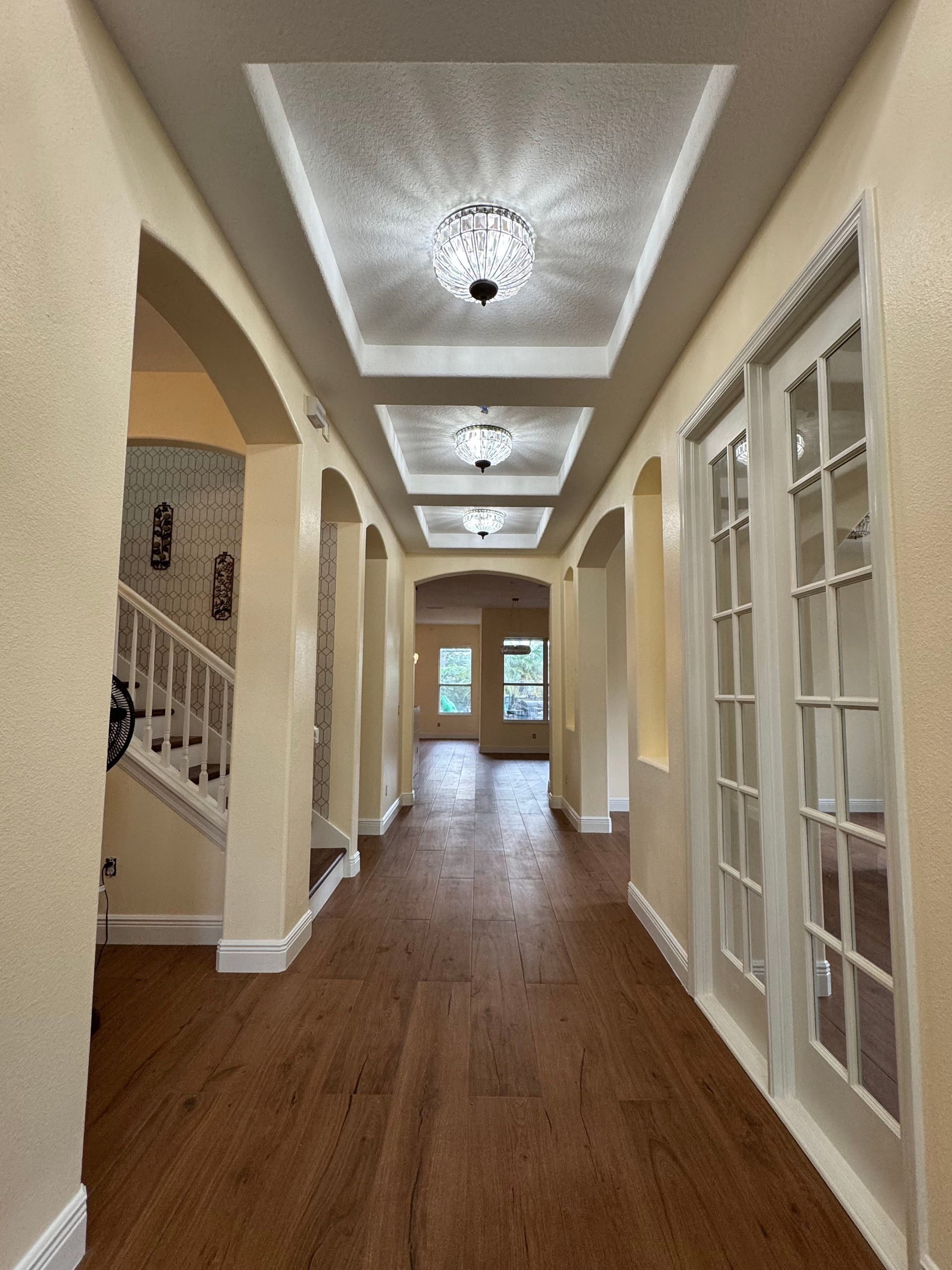 A long residential hallway with wood floors, cream walls, arched entryways, and three recessed ceiling light fixtures.