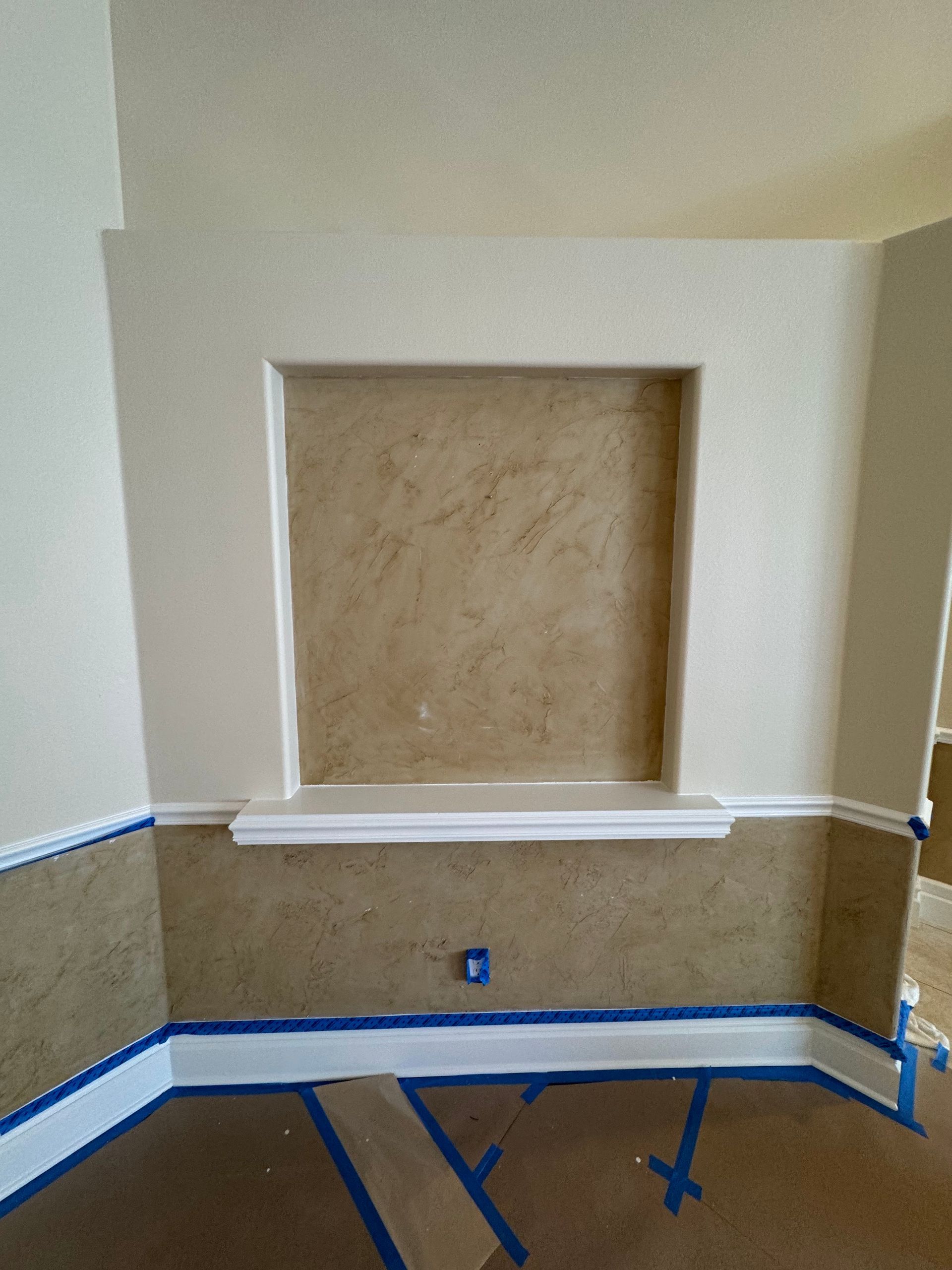 A wall niche with a beige textured back, white trim, and lower walls taped with blue painter's tape for painting.