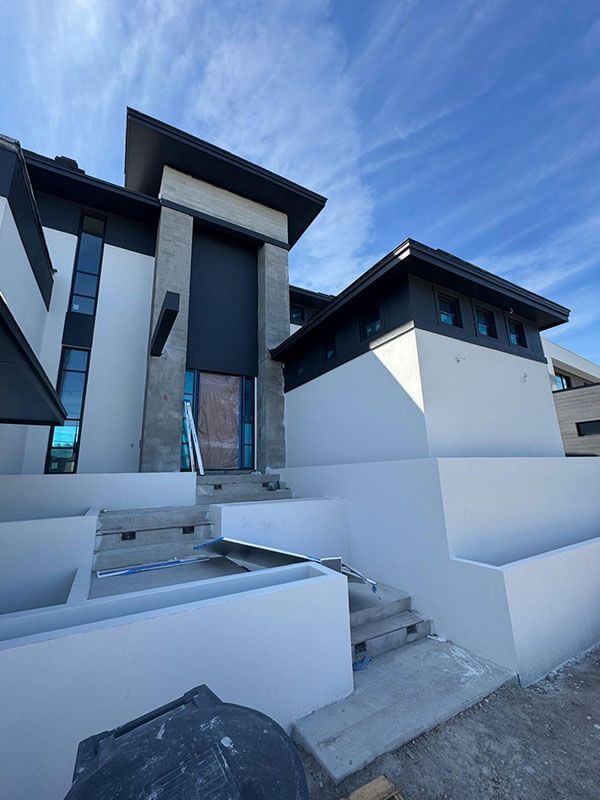 Modern house exterior with white walls, dark gray trim, stone entryway pillars, and concrete stairs under a blue sky.