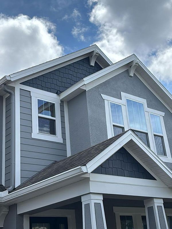 A two-story house exterior featuring blue shingle accents, grey siding, white trim, and a porch under a cloudy sky.