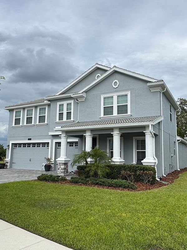 A two-story grey residential house with a front porch, white columns, and a garage under a cloudy sky.