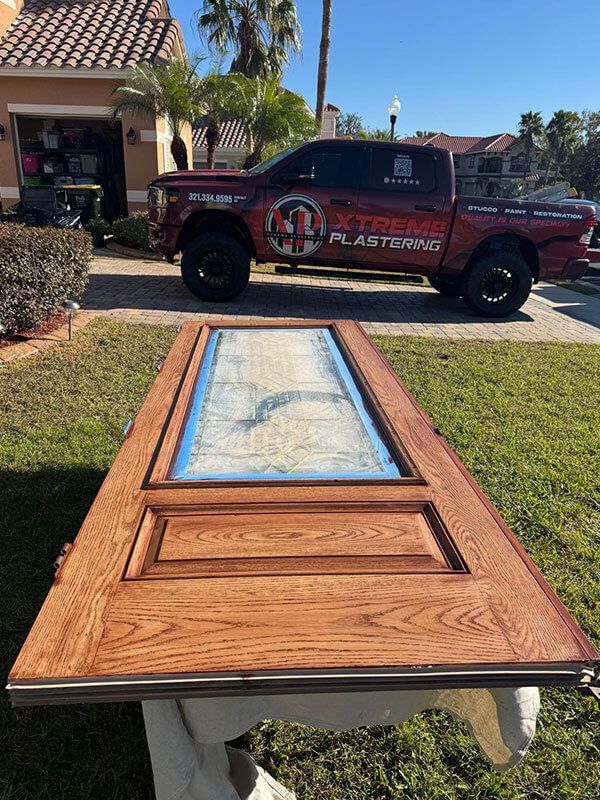 A finished wooden door rests on a stand in a grassy yard, with a dark red Xtreme Plastering truck parked in the background.