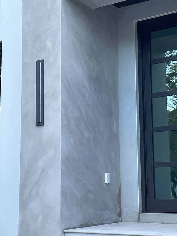 A narrow, dark modern light fixture is mounted on a gray, mottled concrete wall beside a dark-framed glass door.