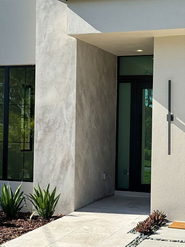 A modern home exterior with a dark glass front door, a textured concrete wall, and a sleek vertical black light fixture.