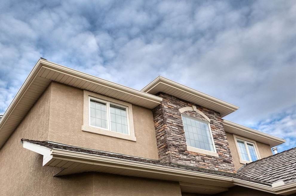 Low-angle view of a modern house with beige stucco walls, stone veneer accents, and white trim against a cloudy sky.