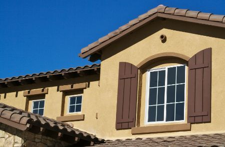 A beige stucco house exterior with brown tile roofing, arched window with shutters, and two smaller square windows.