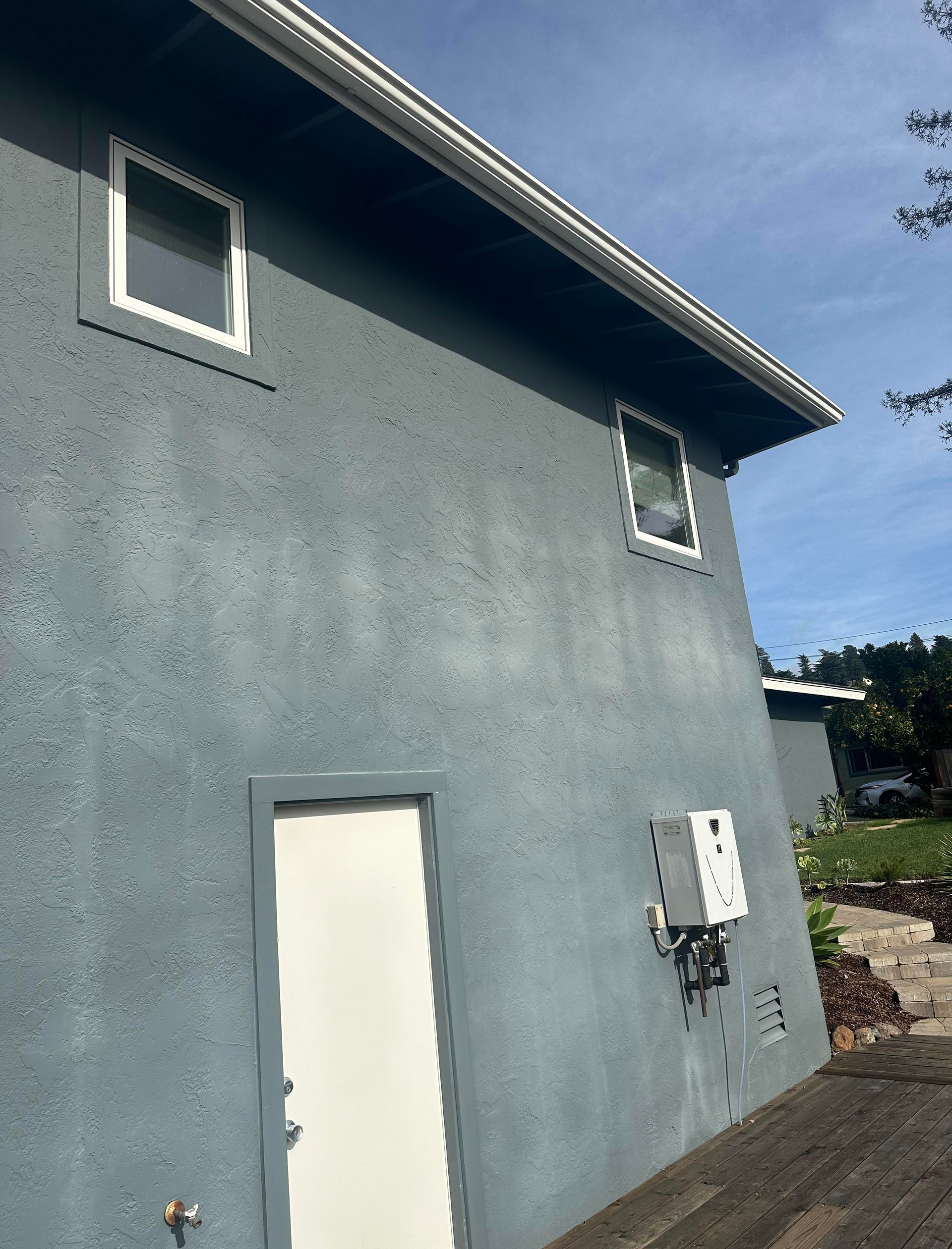 A gray, textured building exterior with two windows, a white door, and an electrical box mounted on the wall.