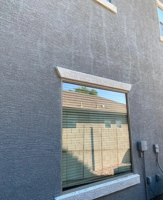 Gray stucco exterior wall with a window featuring light-colored decorative molding and vertical water staining streaks.