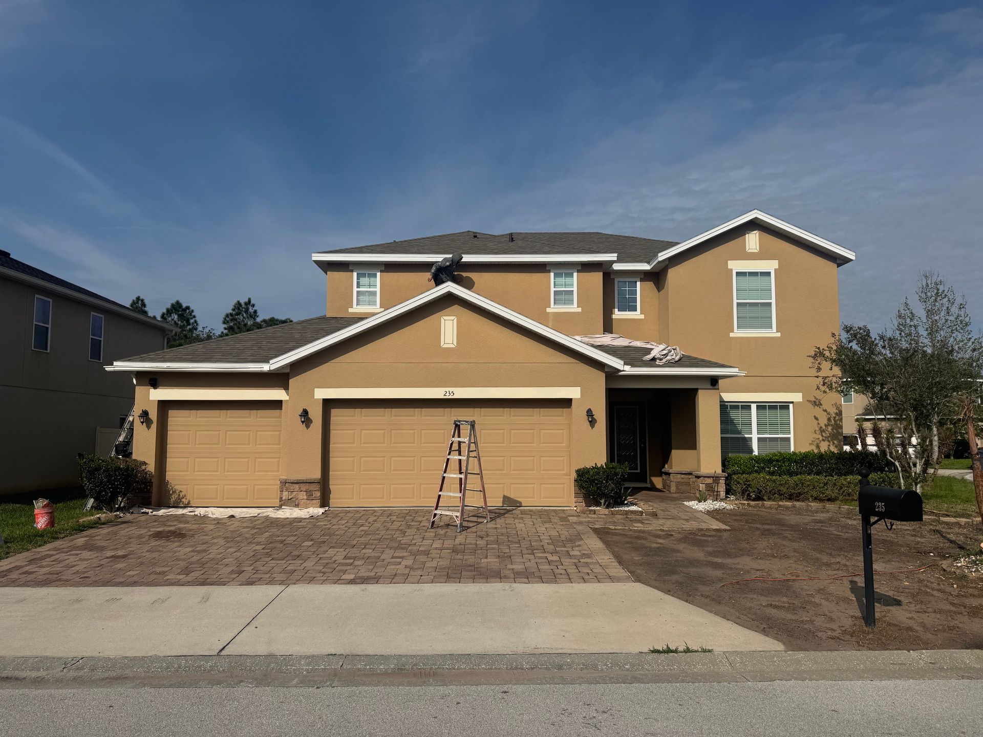 A two-story tan suburban house with a paved driveway and a person working on the roof near a ladder.