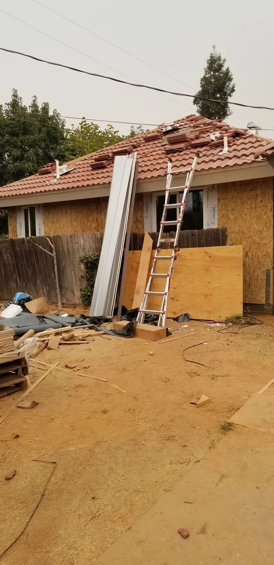 Single-story house with damaged red-tile roof, ladders leaning against it, and debris in a muddy yard.