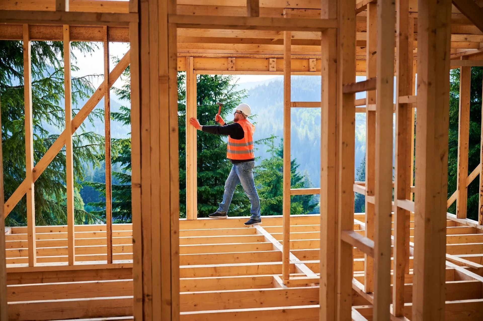 Worker in orange shirt framing a wooden house interior with forest visible through the open structure