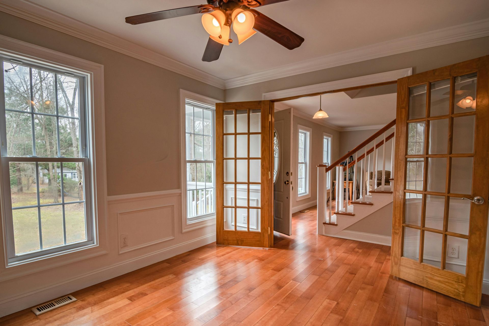 Empty living room with hardwood floors, French doors, staircase, and large windows.