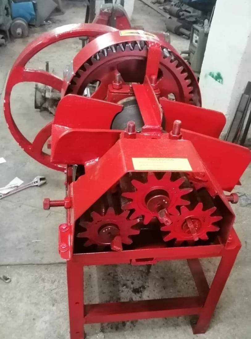 Una máquina industrial de color rojo brillante, accionada por engranajes, probablemente una prensa de jugo de caña de azúcar, se encuentra en el suelo de un taller.