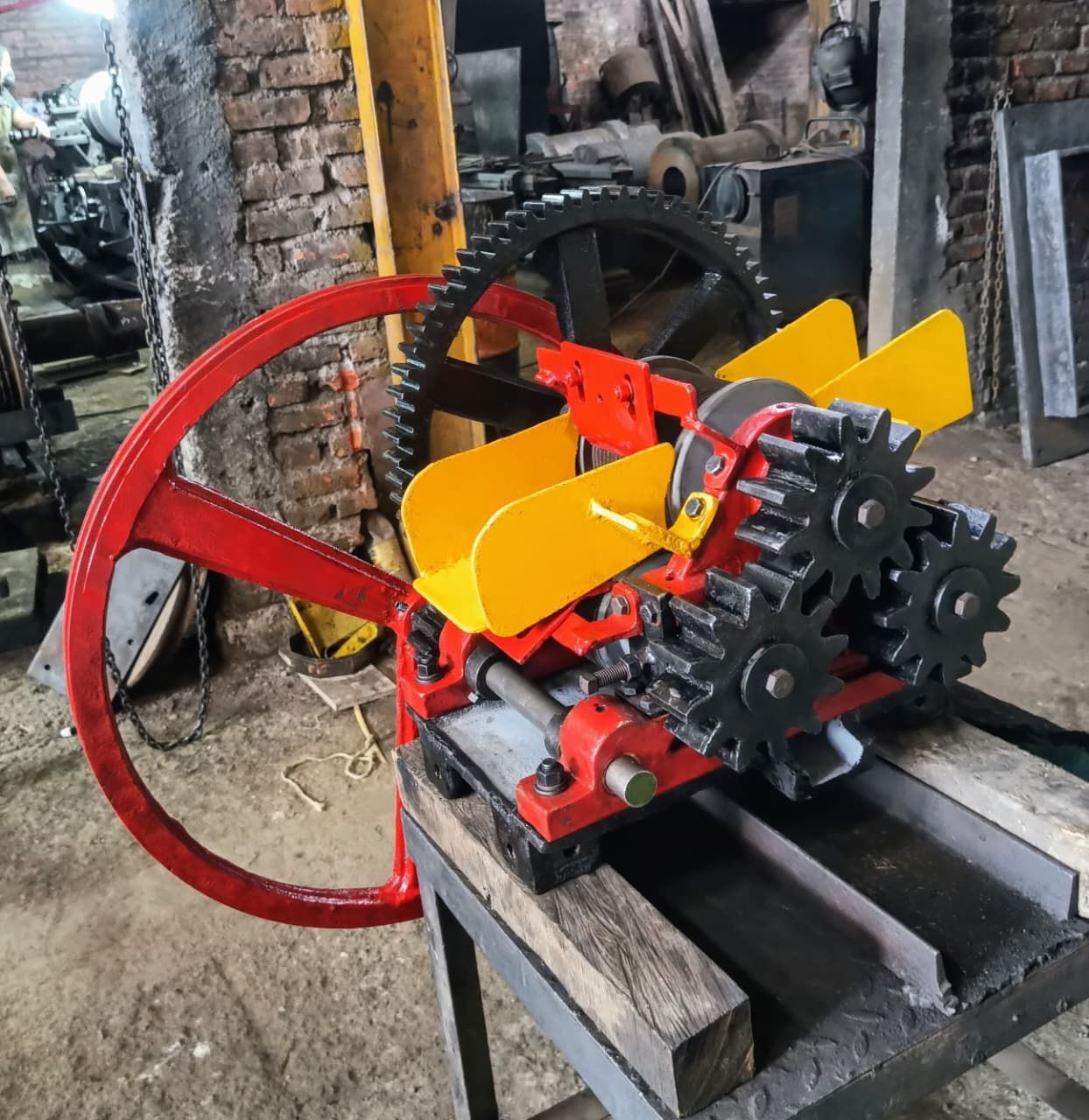 Una máquina metálica roja y amarilla con grandes engranajes y un volante de inercia reposa sobre un banco de trabajo en un taller industrial.