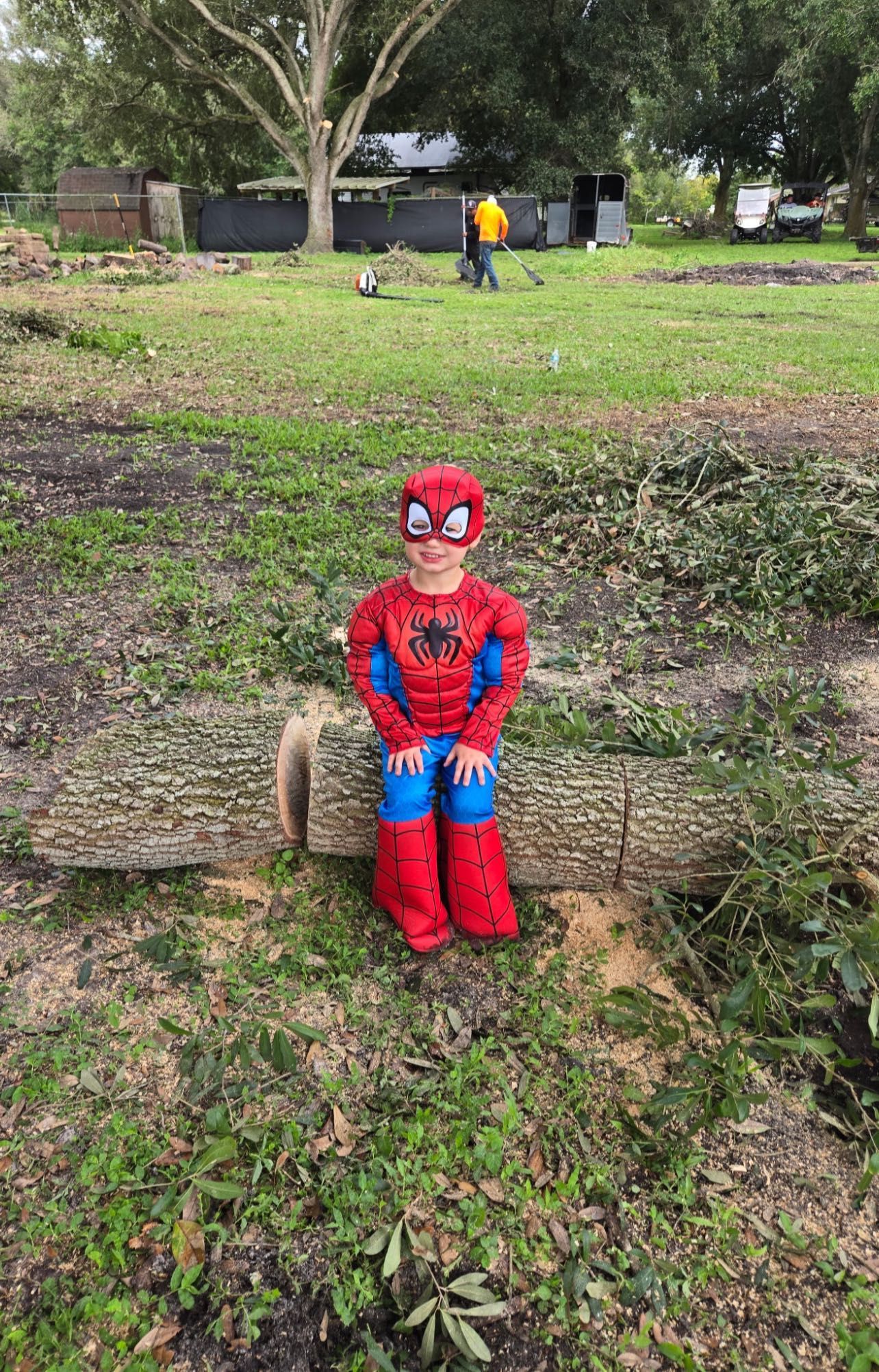 Boy in a Spider-Man costume smiles, sitting on a large log outdoors.