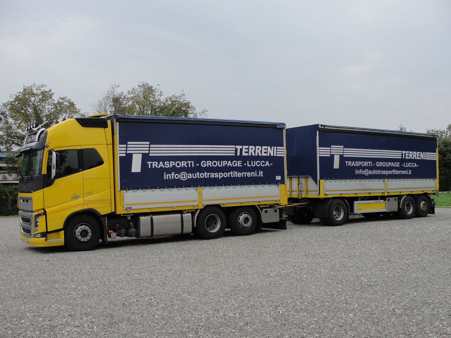 Un camion giallo con rimorchio blu agganciato, parcheggiato su un piazzale ghiaioso sotto un cielo nuvoloso.