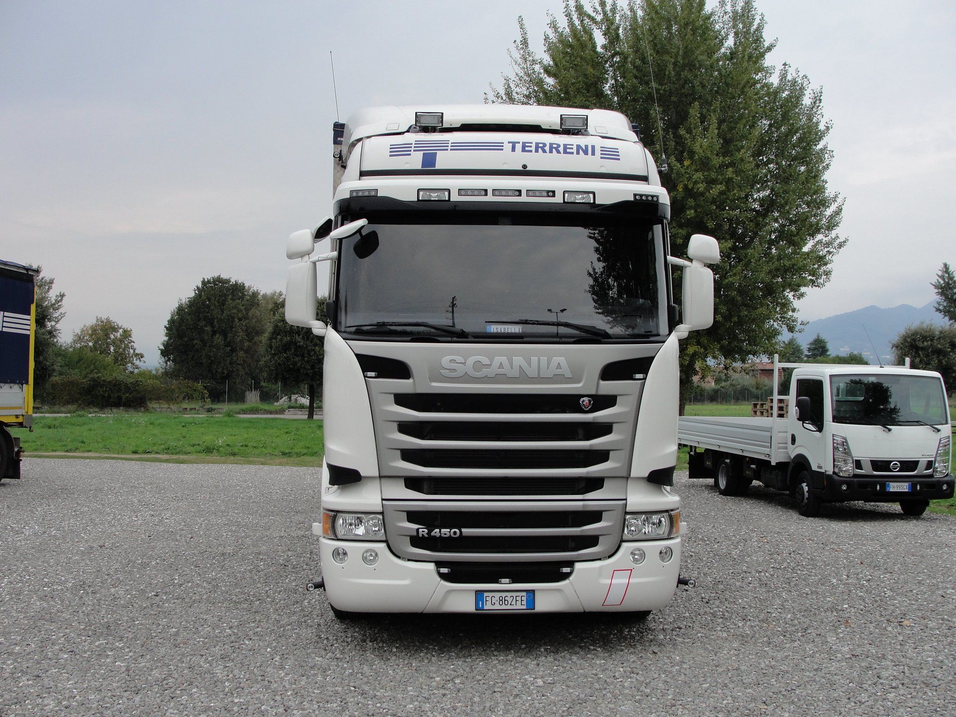 Un semirimorchio Scania bianco parcheggiato su un piazzale ghiaioso, con un camioncino più piccolo a pianale visibile sulla destra.