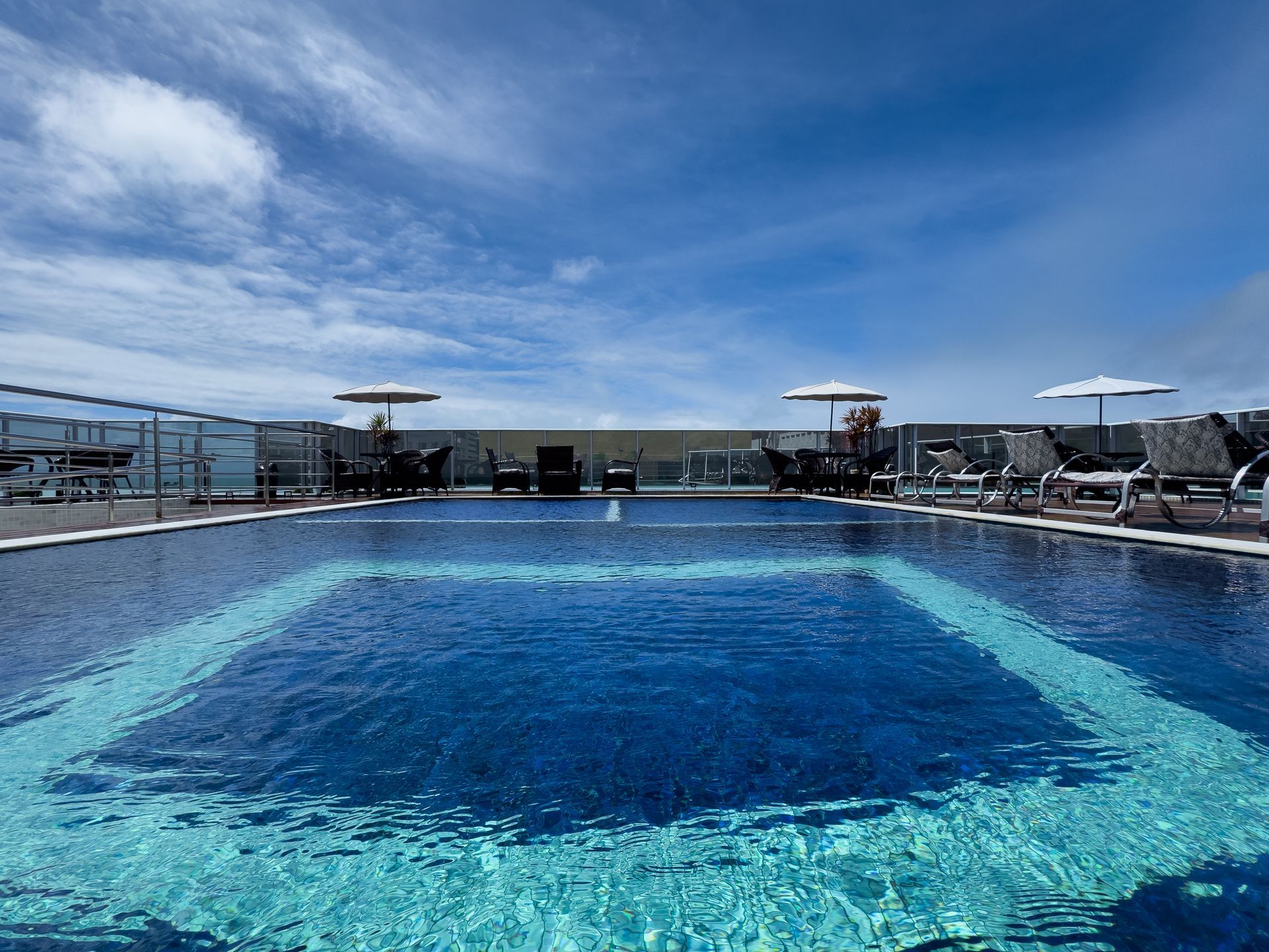 Piscina na cobertura com água azul, guarda-sóis, espreguiçadeiras e um céu azul brilhante.