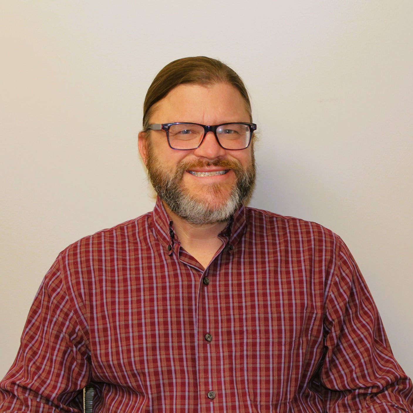 A man with a beard and glasses is wearing a red plaid shirt.