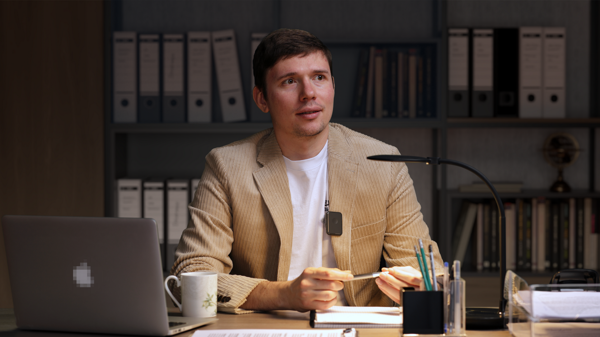 A person in a beige blazer sits at a desk with a laptop, lamp, and office shelves in a dimly lit office setting.