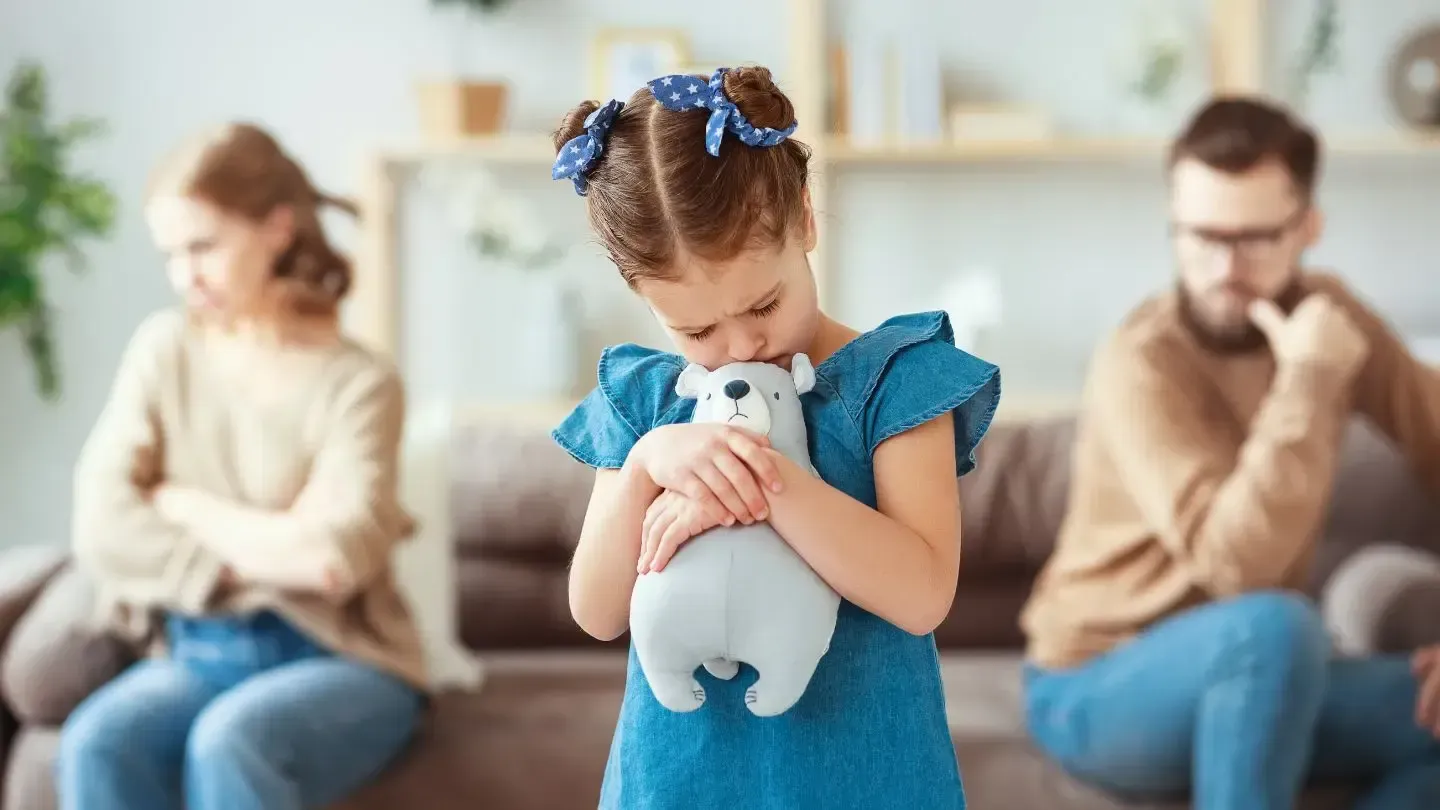 A little girl is holding a teddy bear in front of her parents who are sitting on a couch.