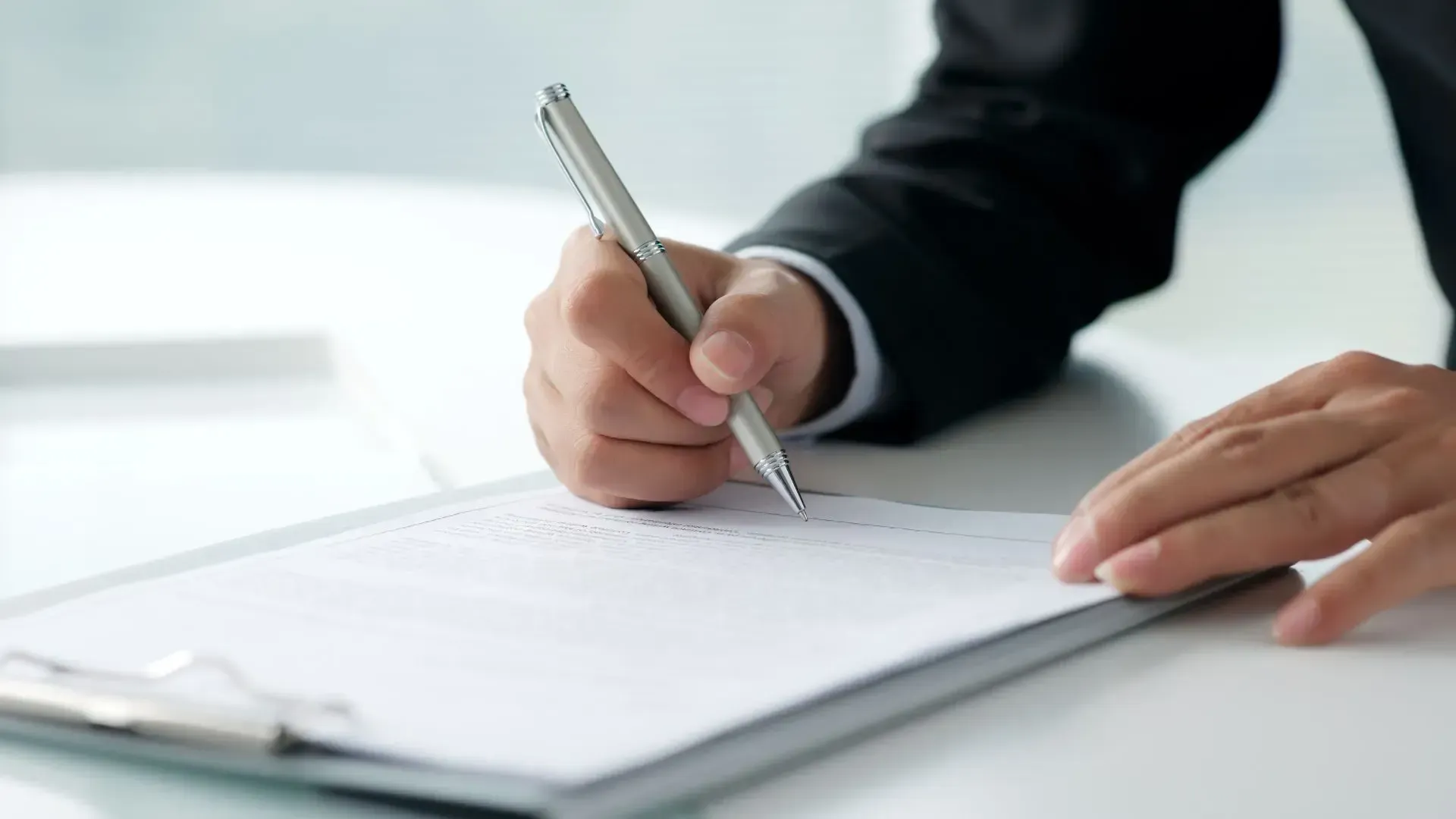 Person in a suit writing on a clipboard with a silver pen; close-up shot.
