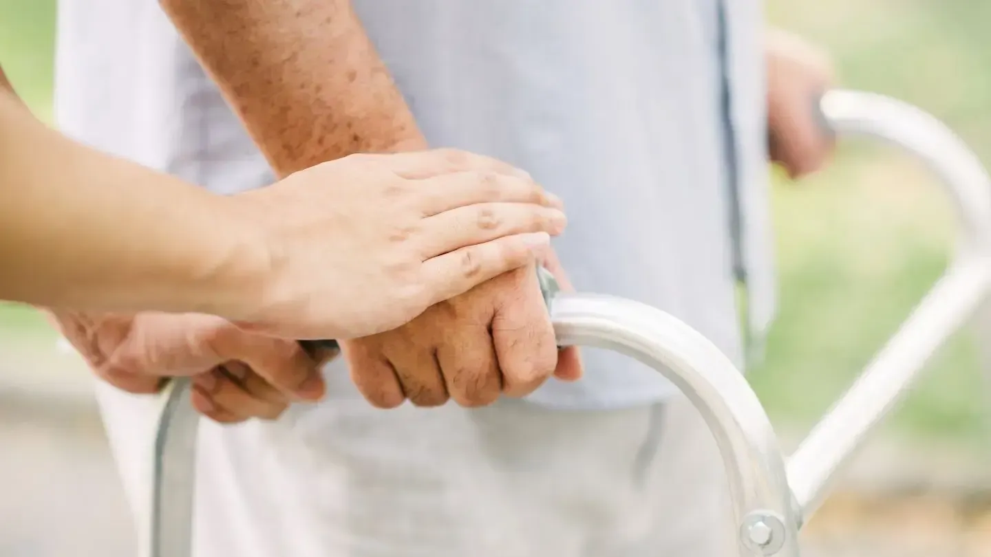 A person using a walker is assisted by another person. A hand rests on the person's arm, and the walker is in the foreground.
