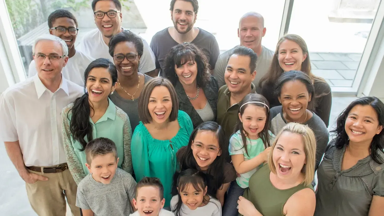Group of diverse people, adults and children, smiling brightly, gathered indoors, with a bright window in the background.