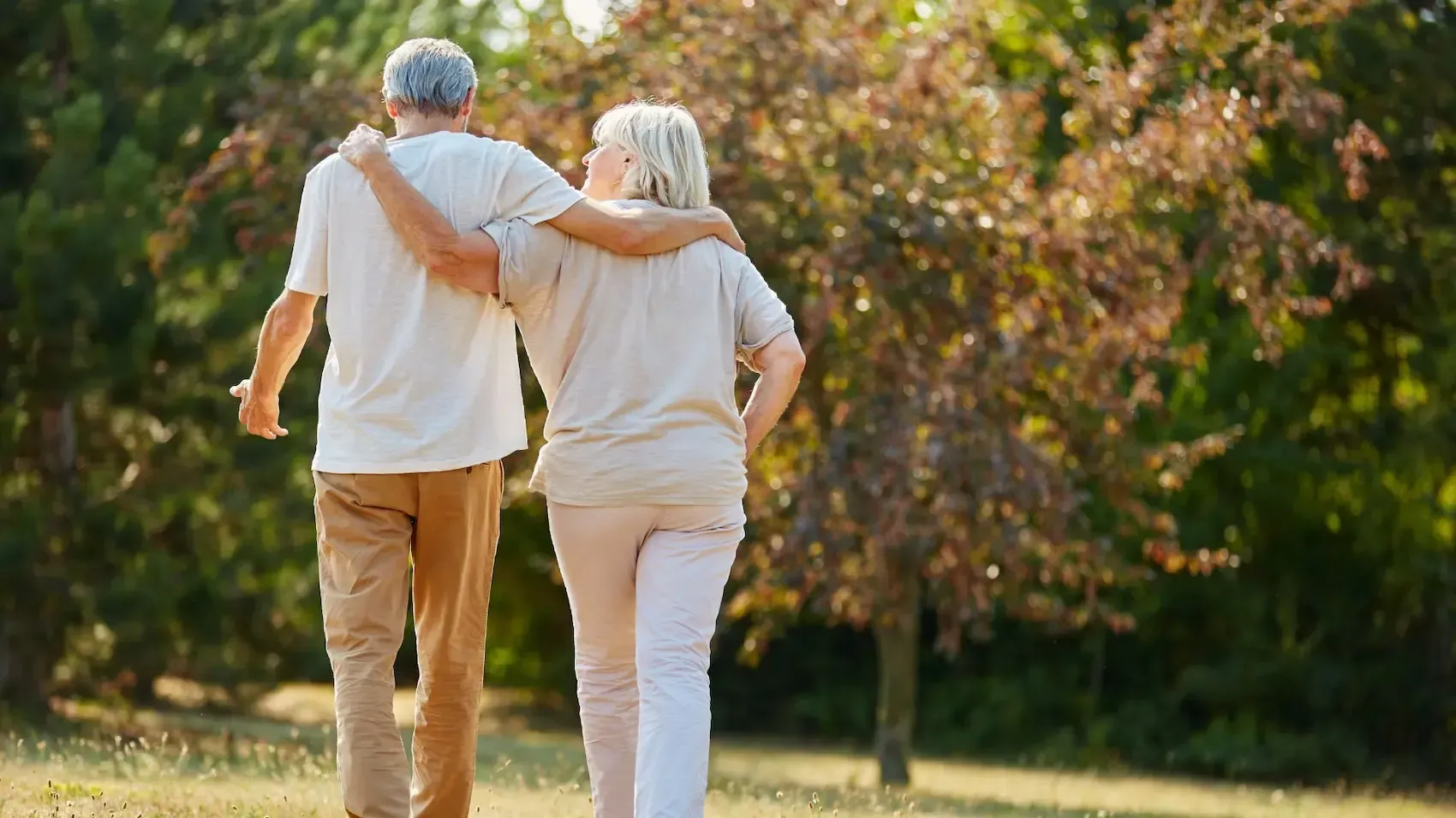 Older couple with arms around each other walking in a sunny park.