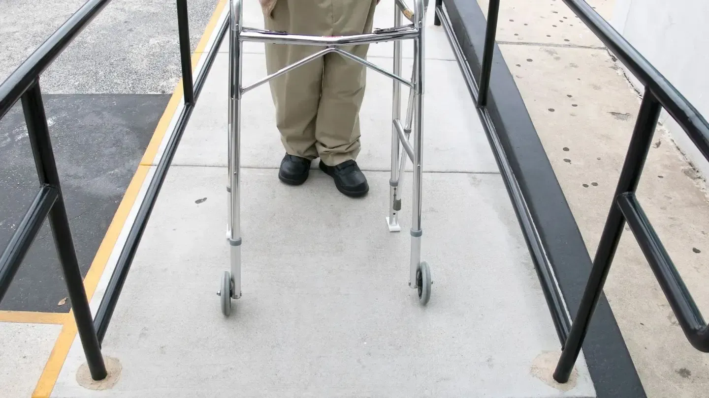 Person using a walker on a concrete ramp with black handrails, outdoors.