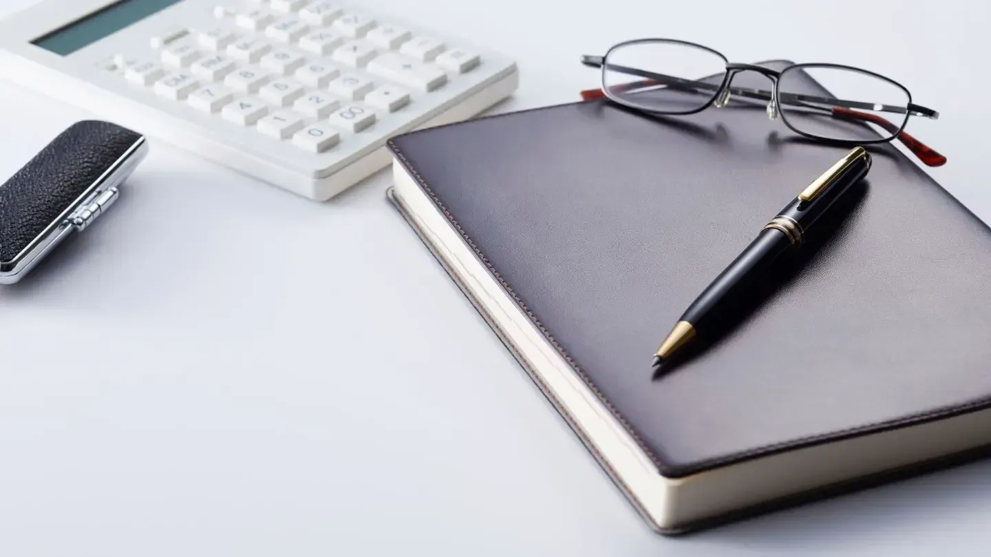 Calculator, journal with pen, glasses, and lighter on a white desk.