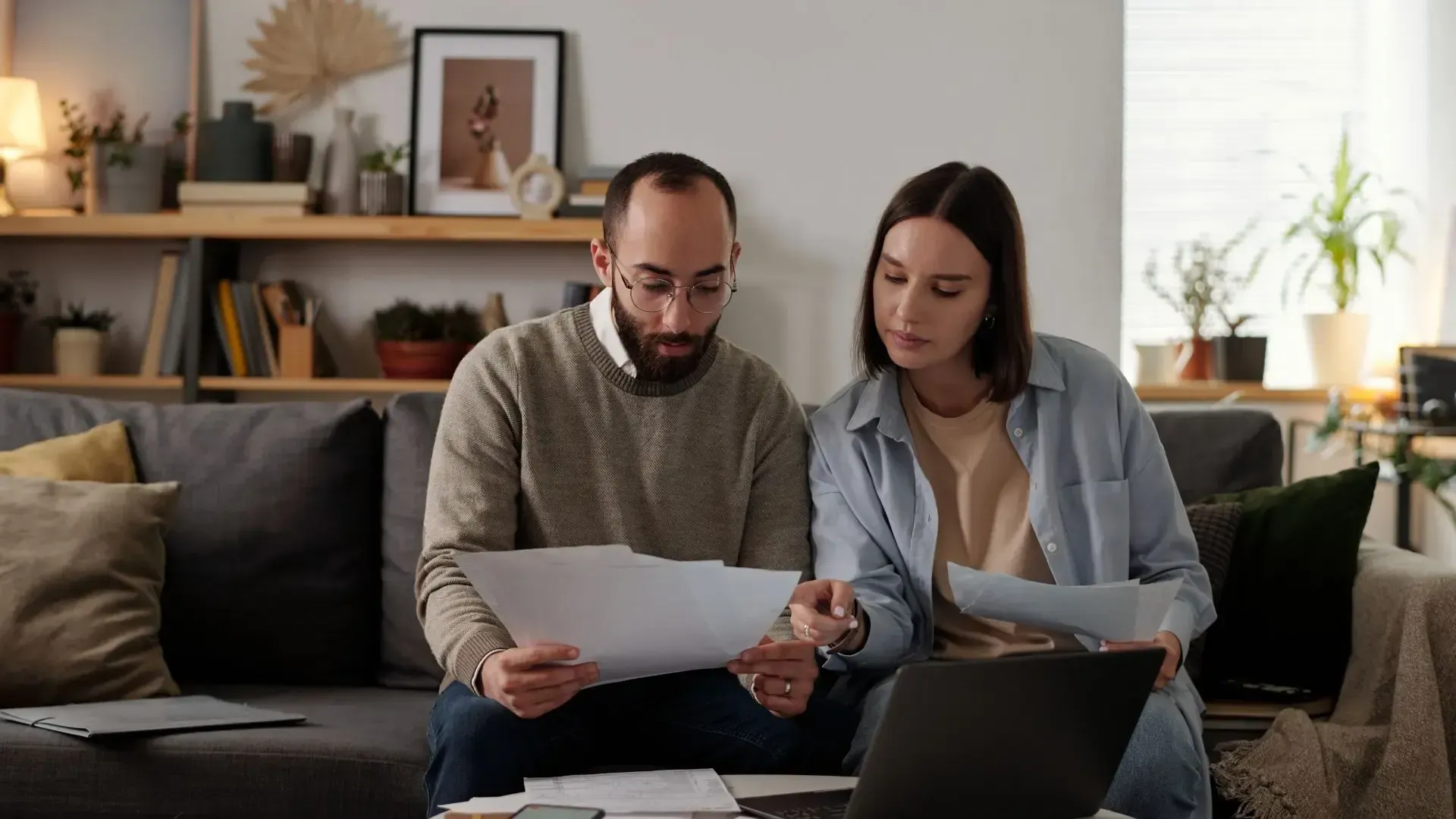 Couple reviewing papers together on a couch with a laptop, likely budgeting or doing finances.