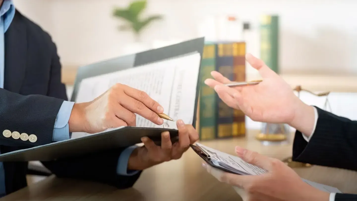 Two people in suits at a desk: one points to documents in a binder, the other reaches for a pen.