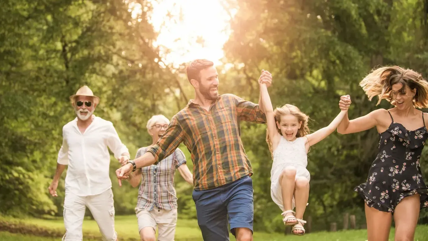 Family holding hands, running in a park. Sunlit background, joyful expressions, grandparents, parents, child.