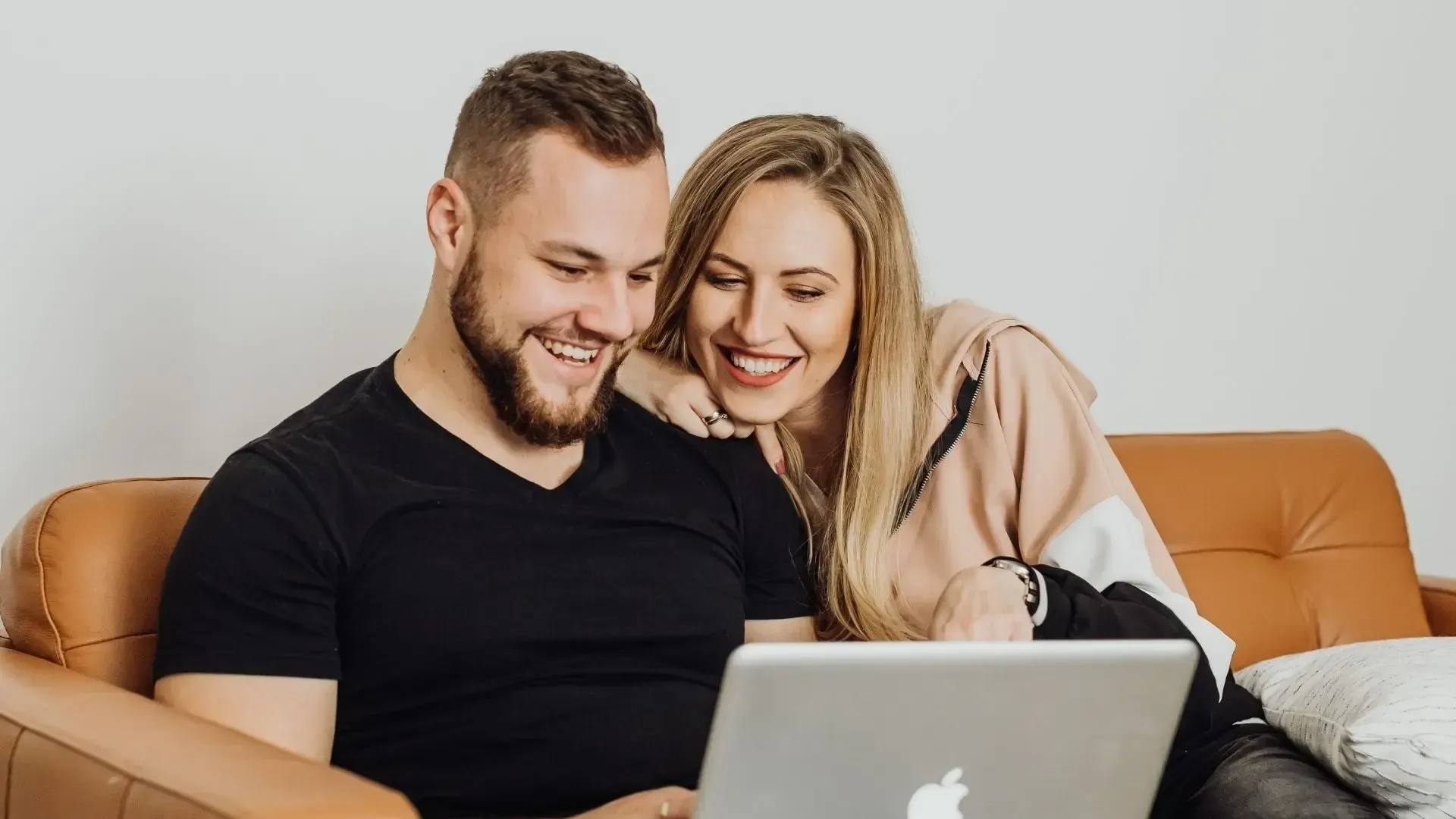Couple smiles while looking at a laptop together on a brown couch.