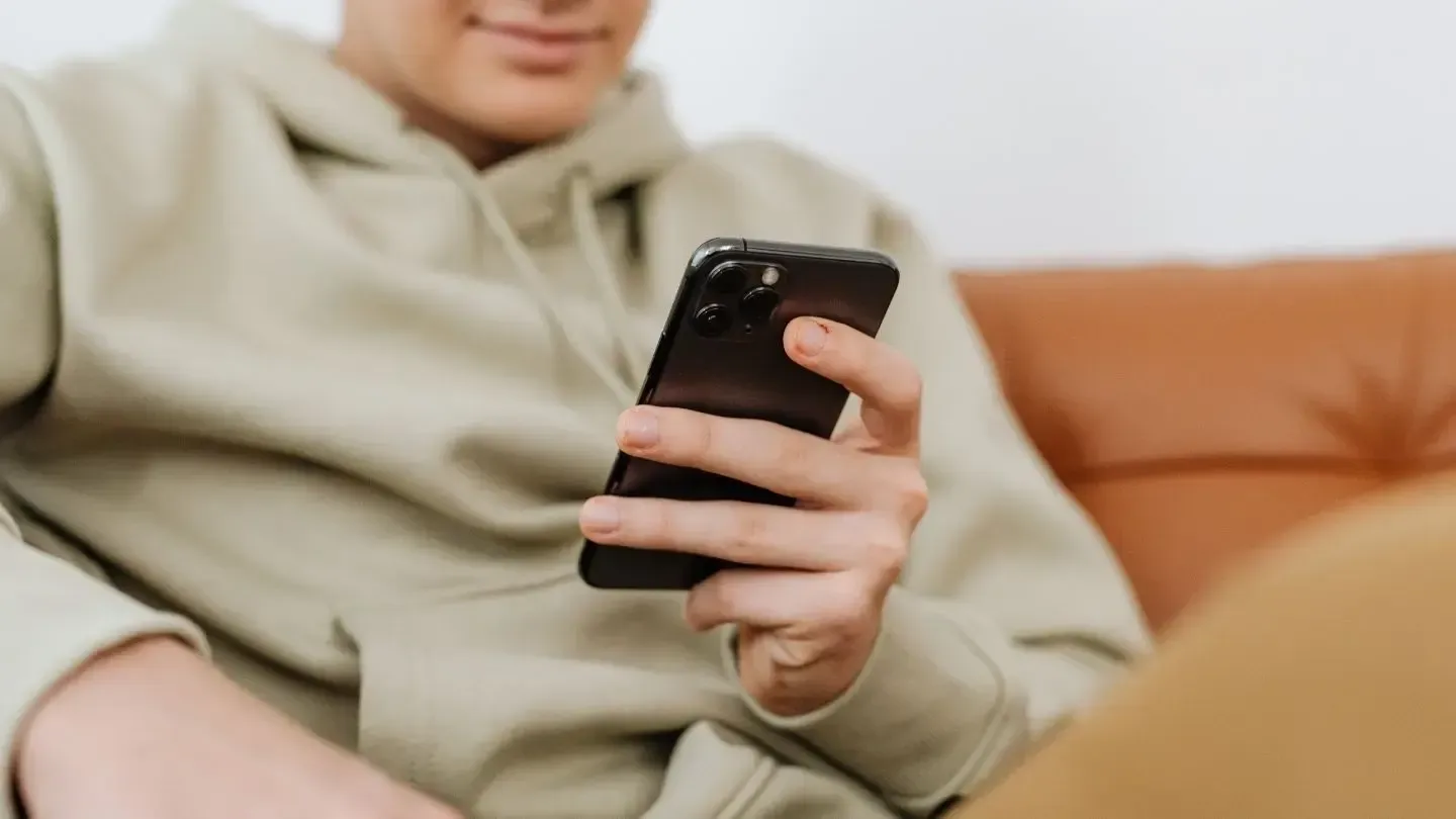 Person in green hoodie, holding a black smartphone, smiling, sitting on a brown couch.