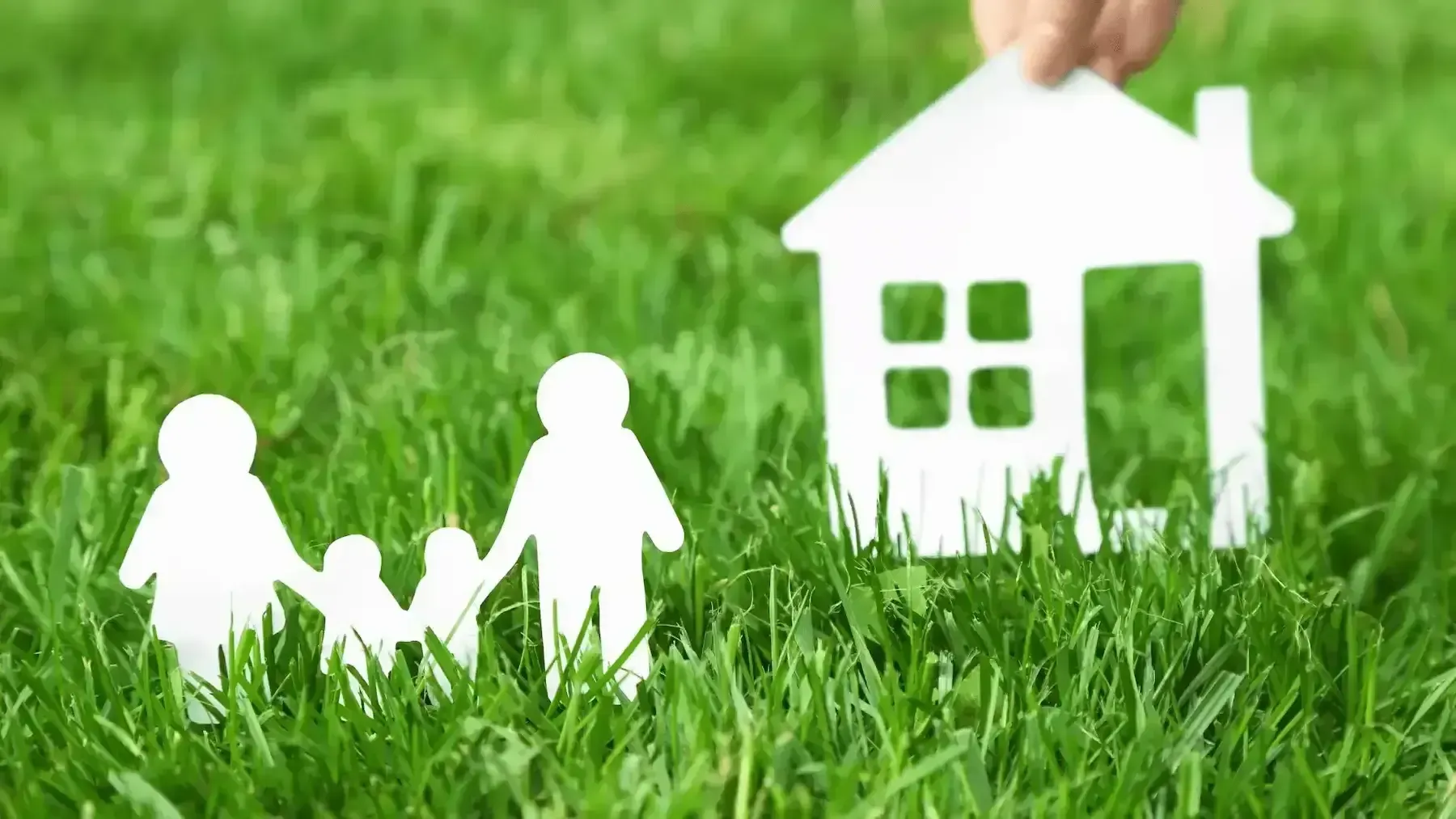 A person is holding a paper house and a paper family in the grass.