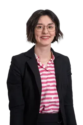 Woman wearing glasses, smiling, in a black blazer and pink striped shirt against a white background.