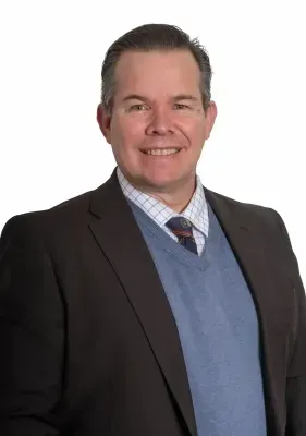 Man in blazer, sweater, and tie smiles. Medium shot, against white background.