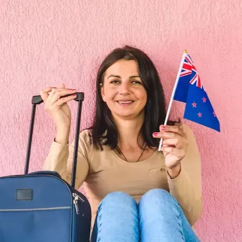 Woman holding a New Zealand flag and suitcase, smiling in front of a pink background.