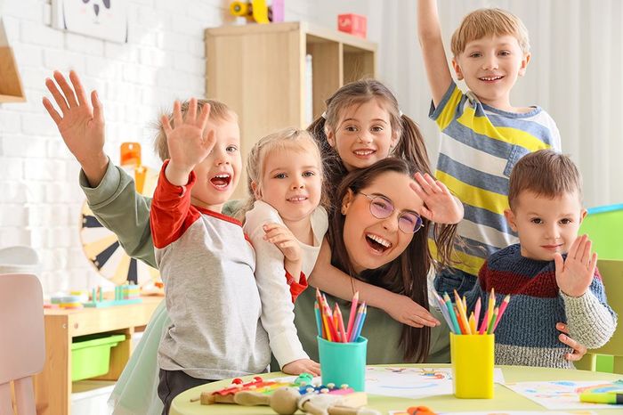 A group of children and a teacher smile and wave, surrounded by colorful art supplies at a table in a classroom.
