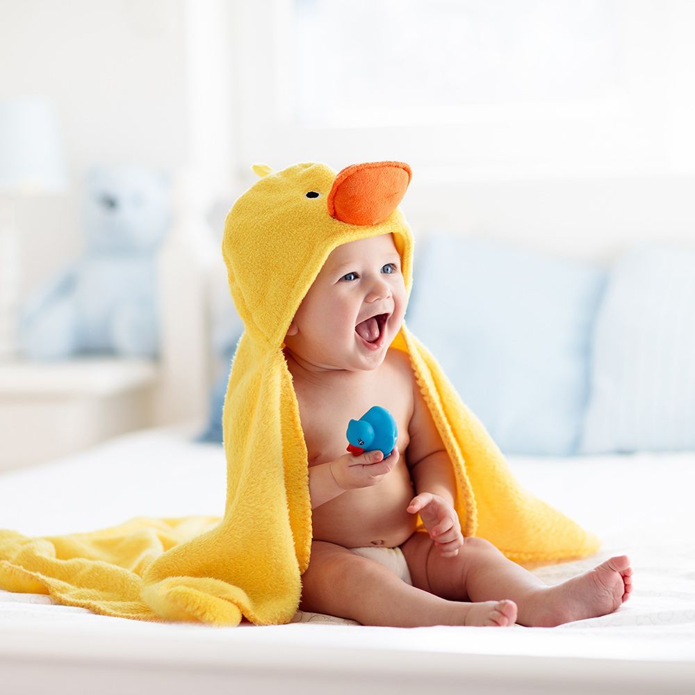 Smiling baby, wrapped in a yellow duck-themed towel, holding a toy in a bright bedroom.