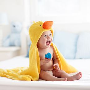 Smiling baby, wrapped in a yellow duck-themed towel, holding a toy in a bright bedroom.