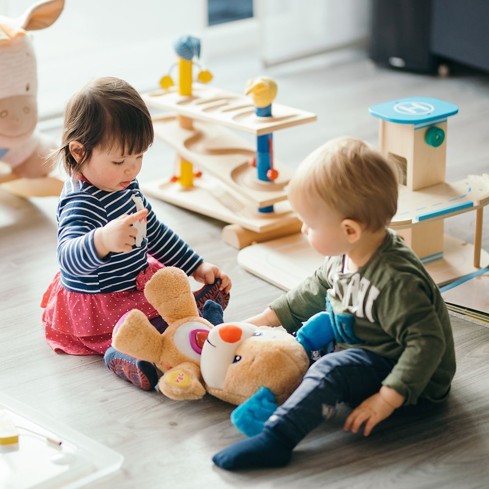 Two toddlers playing on a wooden floor with toys, including a plush bear and wooden car ramp.