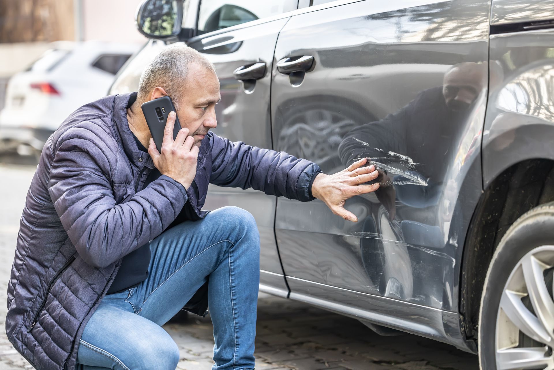 Man on phone examining car with damage. Gray car, blue jacket, outdoors.